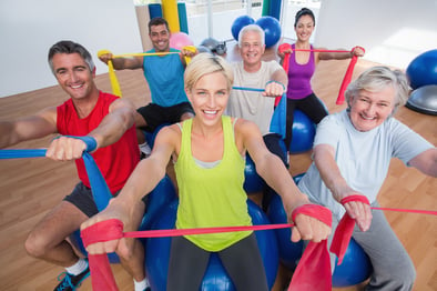 Portrait of happy men and women on fitness balls exercising with resistance bands in gym class-1